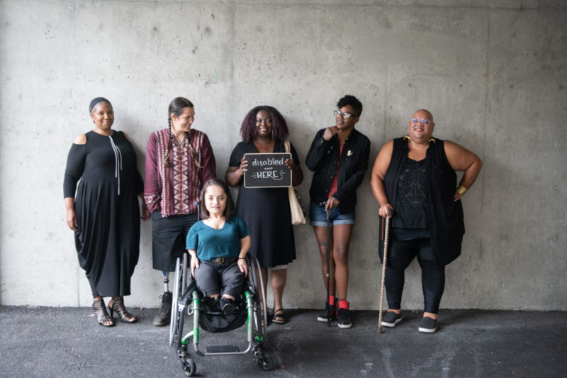Six disabled people of color smile and pose in front of a concrete wall. Five people stand in the back, with the Black woman in the center holding up a chalkboard sign reading 