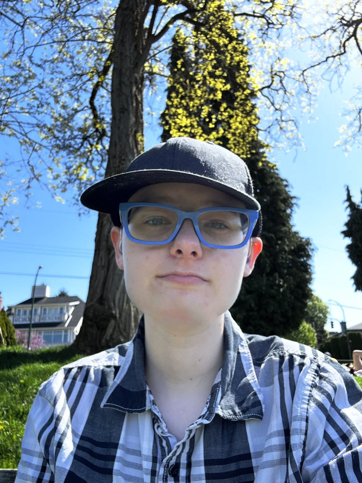 Casey, outside on a sunny day, wearing blue glasses, a ball cap, and a plaid shirt.